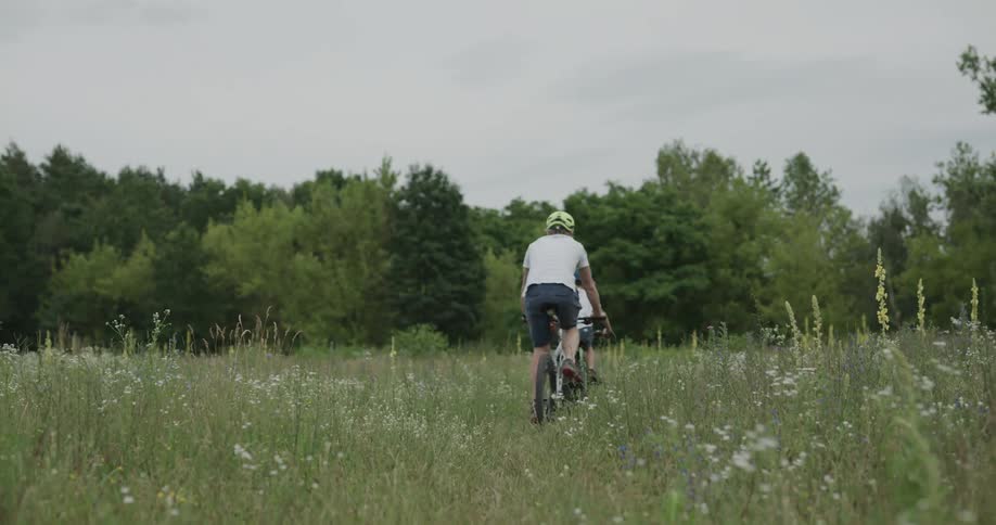 Two kids ride their bikes through tall grass towards the trees
