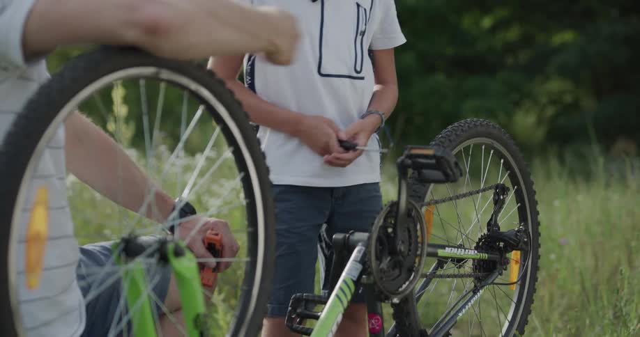 Dad and son wear helmets as they fix a bicycle wheel outdoors next to trees