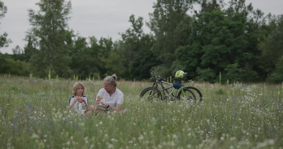 A father and son sit in a field and eat while taking a break from bike riding