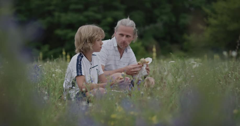 A father and son sit in a field of flowers and eat and the dad laughs as he wipes food off his sons face