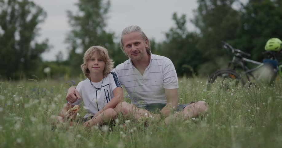 Father and son sitting on the green grass on a field near bicycles looking at the camera