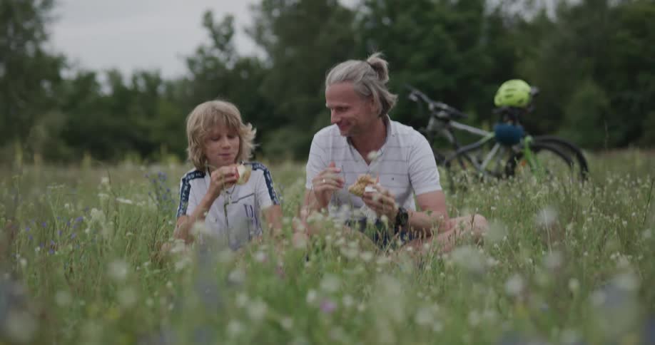 Father and son sharing a snack after cycling sitting in the green grass on a field at day
