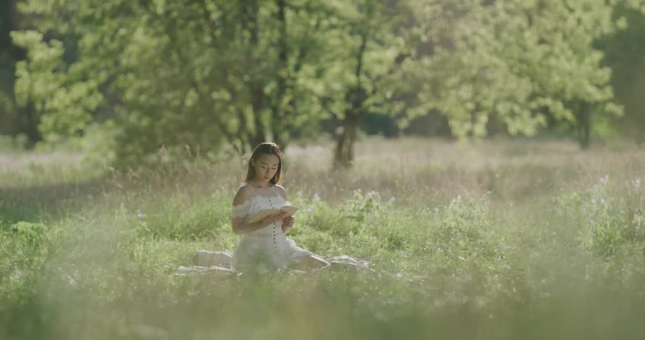 A young asian woman reads a book while sitting on a blanket in a field on a sunny day