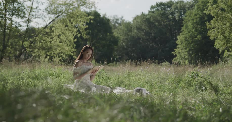Asian woman sitting on a blanket on a meadow near trees reading a book