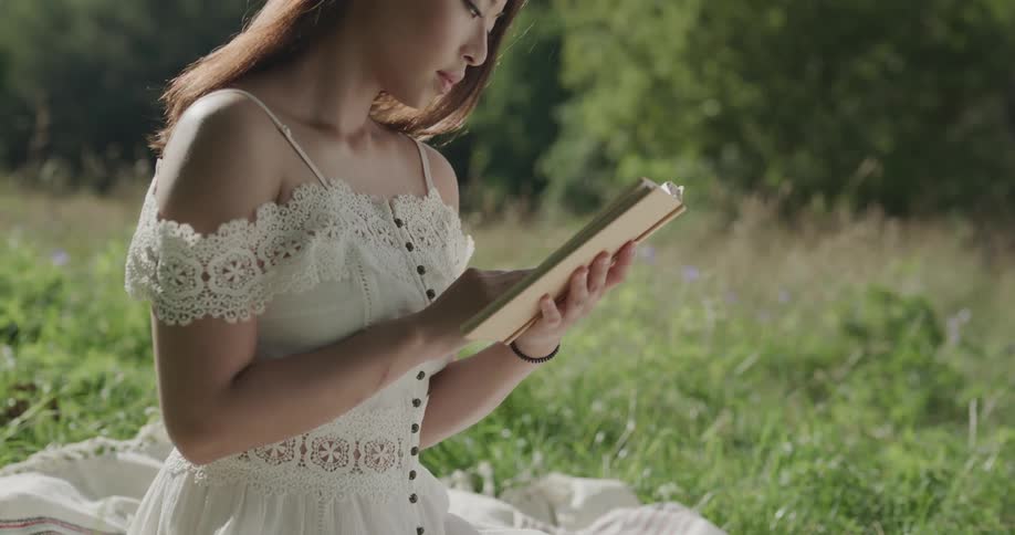 Young woman wearing white dress reading a book on a blanket over the grass at sunset