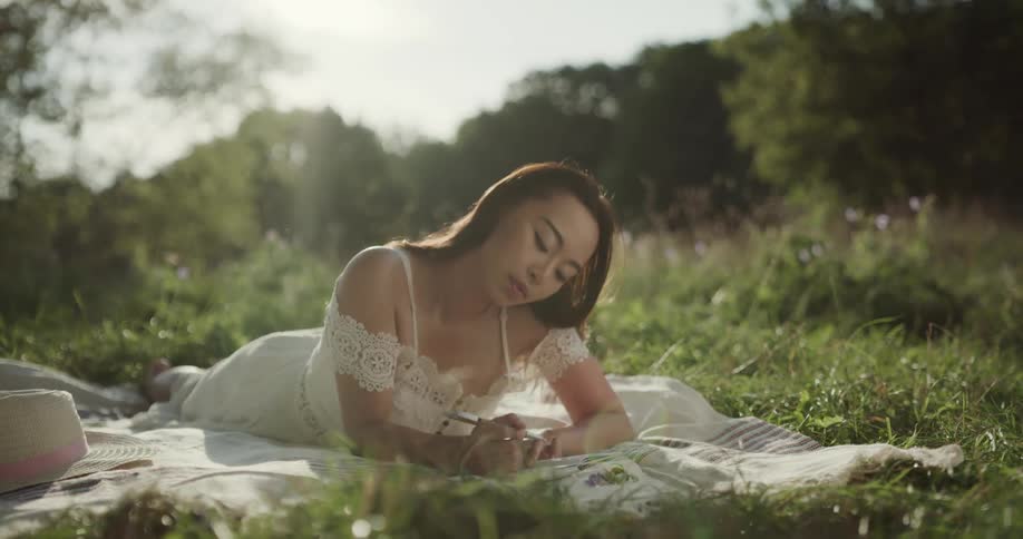 Young woman in a white dress writes a note over a blanket on the grass