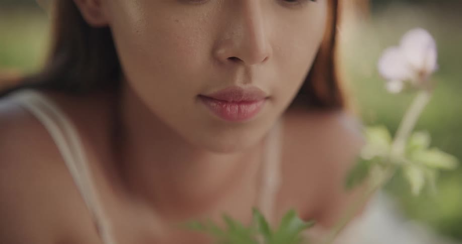 A woman looking and smelling a plant with a purple flower with grass in the background