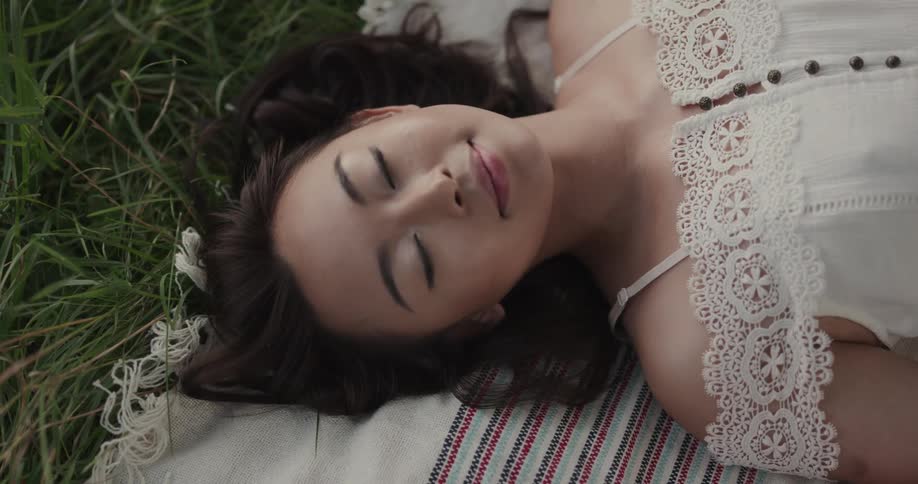 A woman laying down on a blanket on grass with her eyes closed then opening her eyes and staring