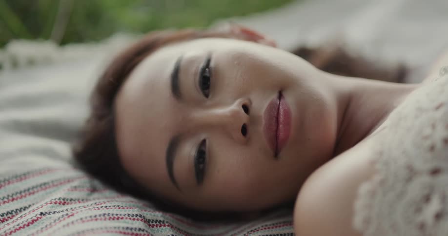 A smiling asian woman laying on a blanket on the grass in a white lace dress