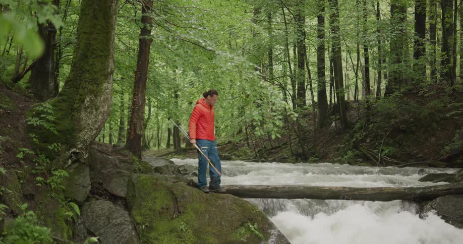 Man wearing orange hoodie walks to river the bank in the woods as the stream is flowing below