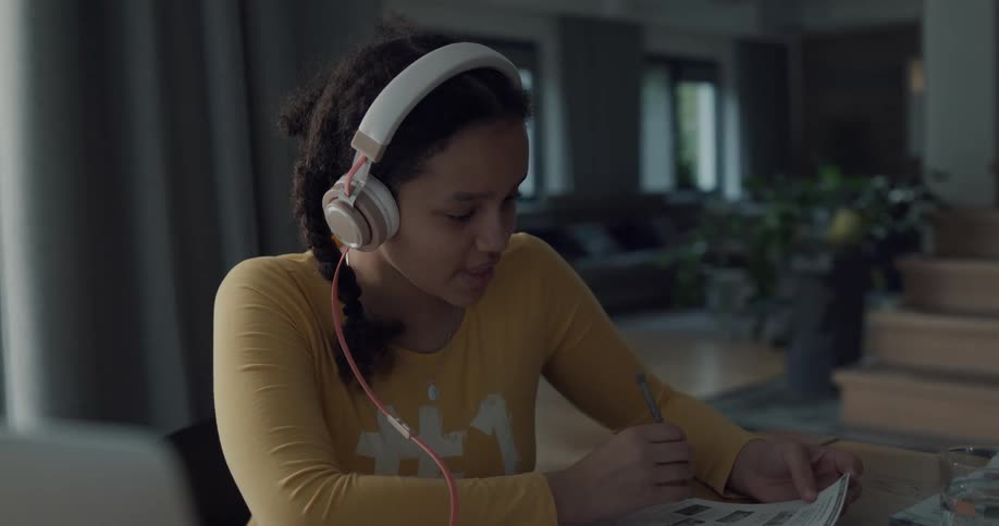 A girl with a pen a book and headphones sitting at a table in front of the laptop