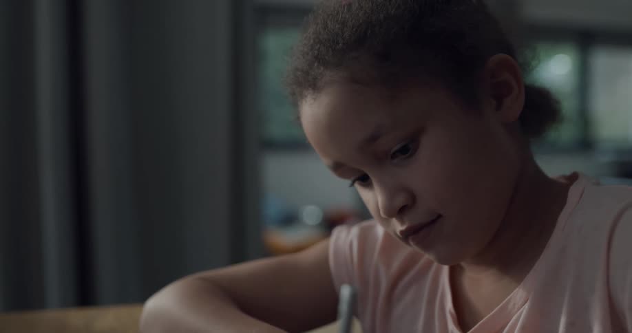 A girl writes in a study book while sitting at a table in a house during the day