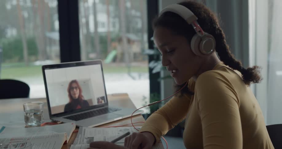 A girl in a room with headphones sitting at a table video calling on a laptop