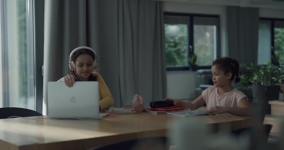 Two girls in a room sitting at a table with one putting on headphones and opening a laptop