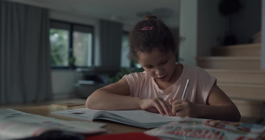 A girl works on homework while sitting at a table by the stairs in her house
