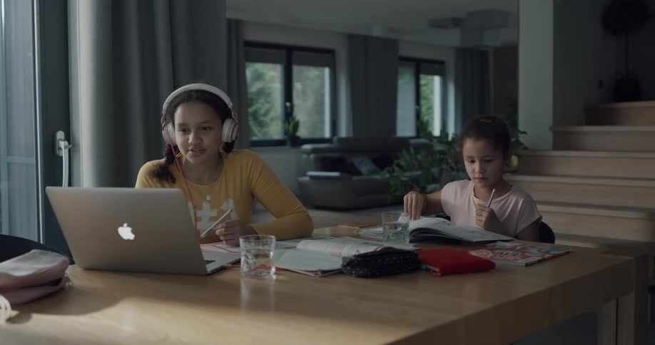 Two girls in a room sitting at a table with one with a headphones on and sitting in front of a laptop