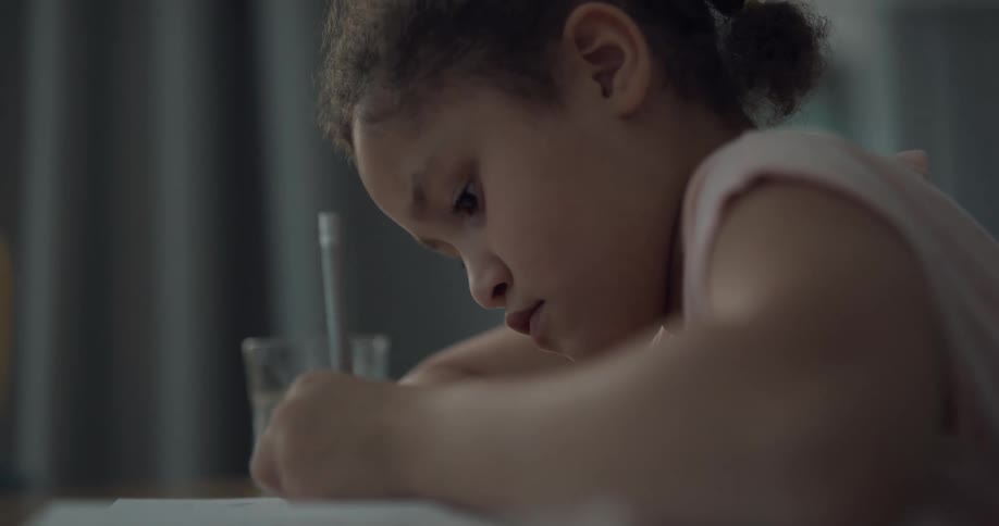 A girl sitting at a table writes in a workbook with a pencil