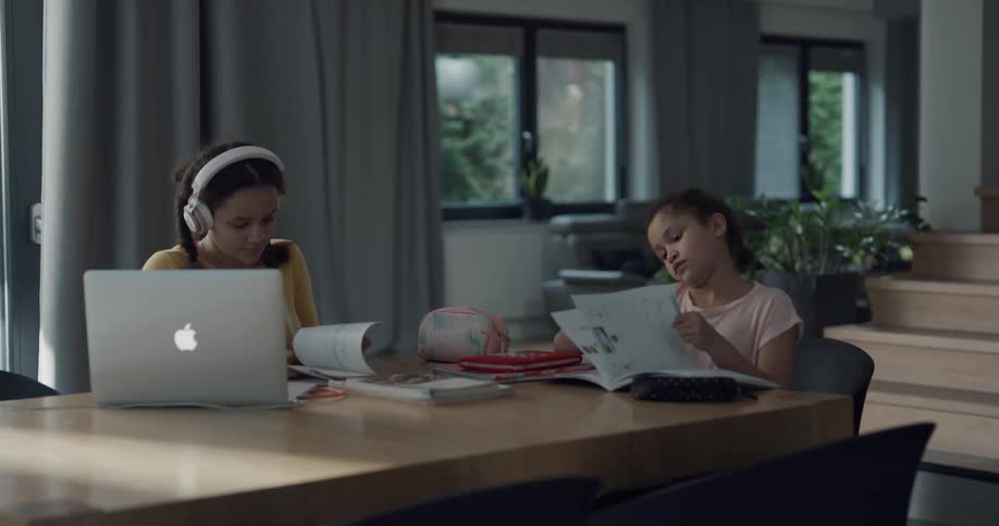 A pair of girls look through workbooks while sitting at a table in a house during the day