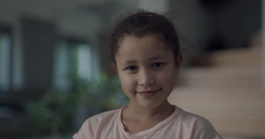 A girl smiles into the camera while standing before a staircase indoors during the day