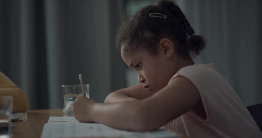 A young girl sits at a table while focused on what she is writing