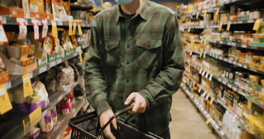 A man wearing a green plaid shirt and jeans places items in his black basket while shopping in a grocery store