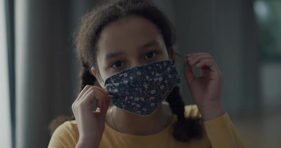 A girl wearing a medical mask looks into the camera while she removes the mask from her face