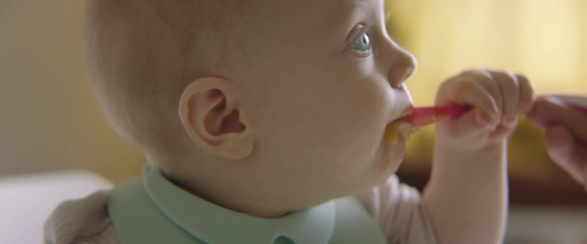 A baby sitting getting fed food from a spoon by another person