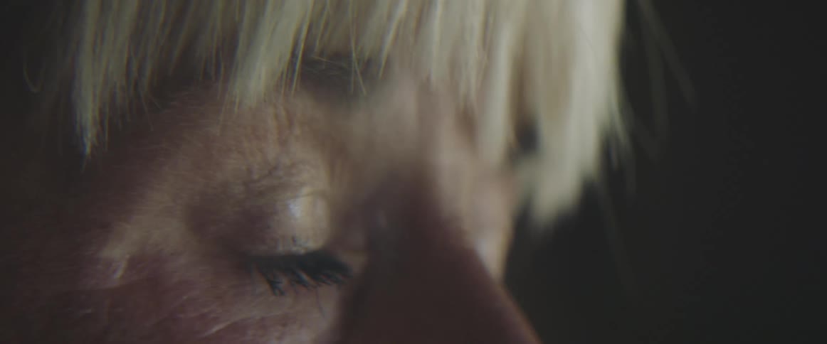 A woman with blonde hair sits in a dark room and blinks away tears