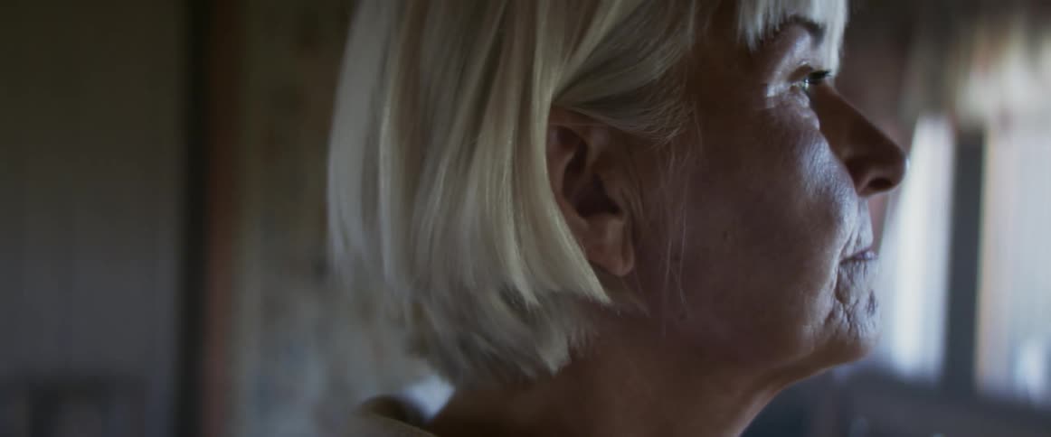 A woman with a short blonde bob haircut stands in a living room with decorative wallpaper