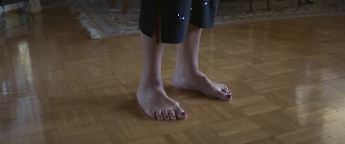 An elderly woman walks in to a room at home and looks out the window