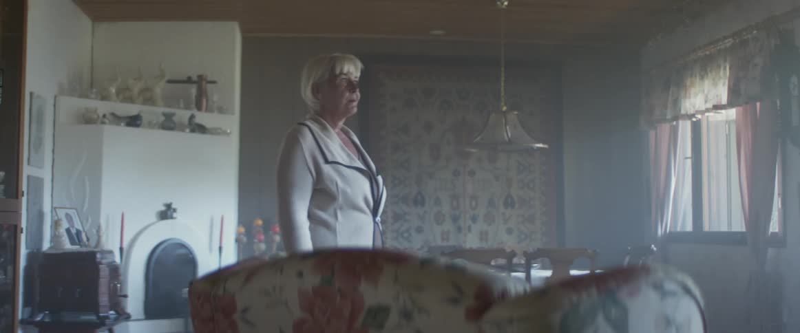 Elderly woman with grey hair walks in a room and looks out the windows