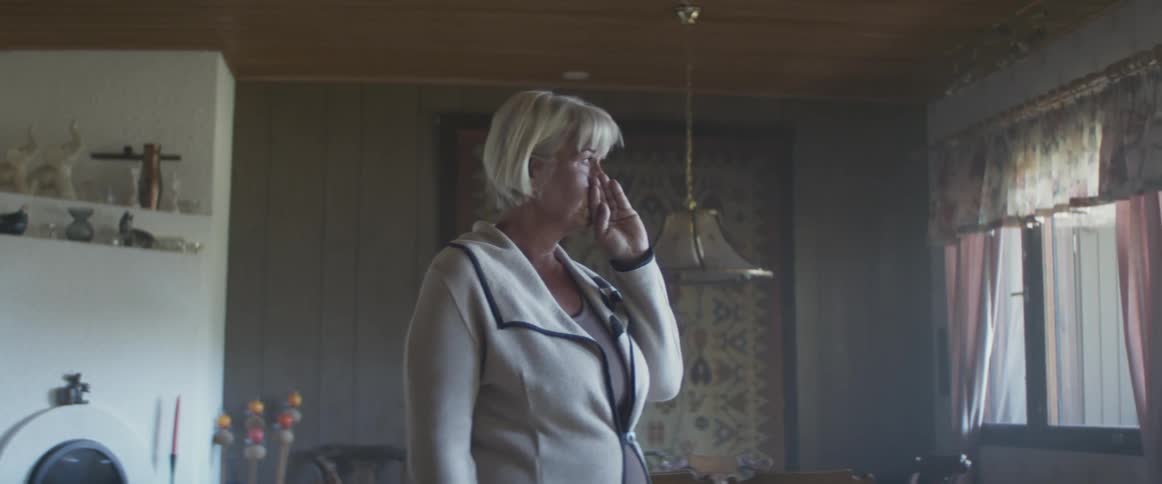 Elderly woman with grey hair stands and wipes tears with hand in dining room