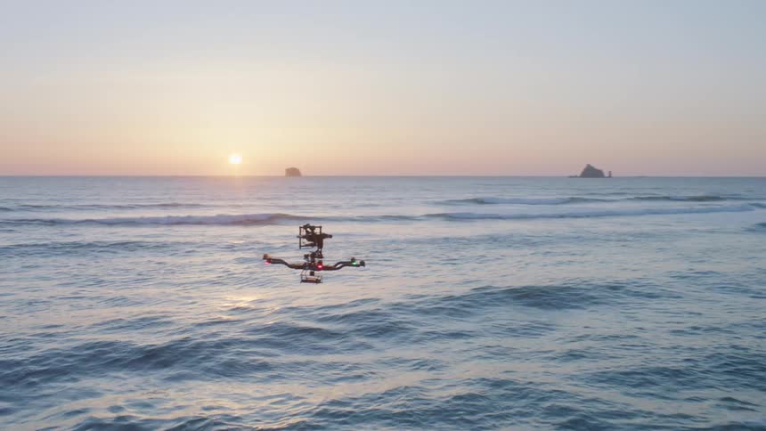 Close flyby of a commercial drone hovering over breaking ocean waves as the sun glows low along the horizon