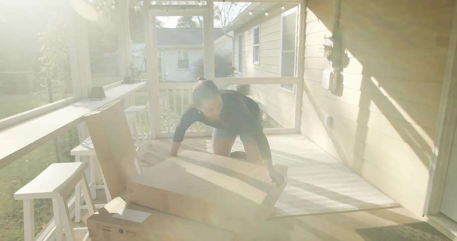 Young woman unboxing furniture from cardboard boxes on the porch at sunset