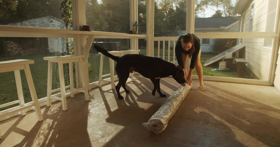 A woman unpacking a rug in the porch at home with her dog at sunset