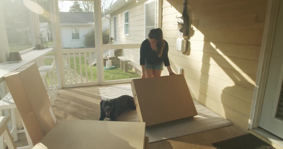 A woman moves a cardboard box to the floor of her porch with a black dog watching her