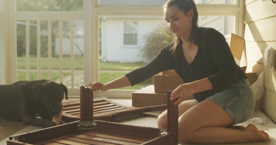 A womans dog sits nearby and chews on a bone as she builds a chair on her screened in porch