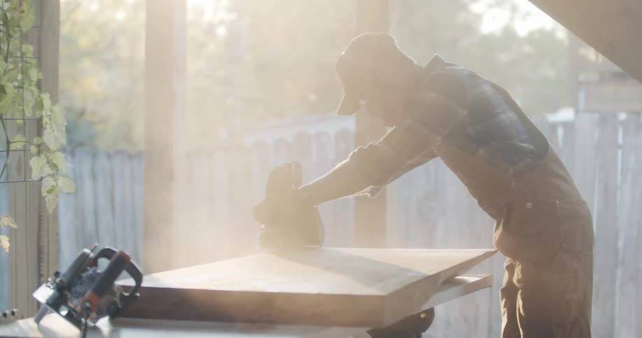 A man uses a power sander on a piece of wood outside and wearing face protection