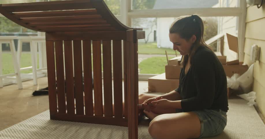 A woman sits on a porch assembling a wooden chair