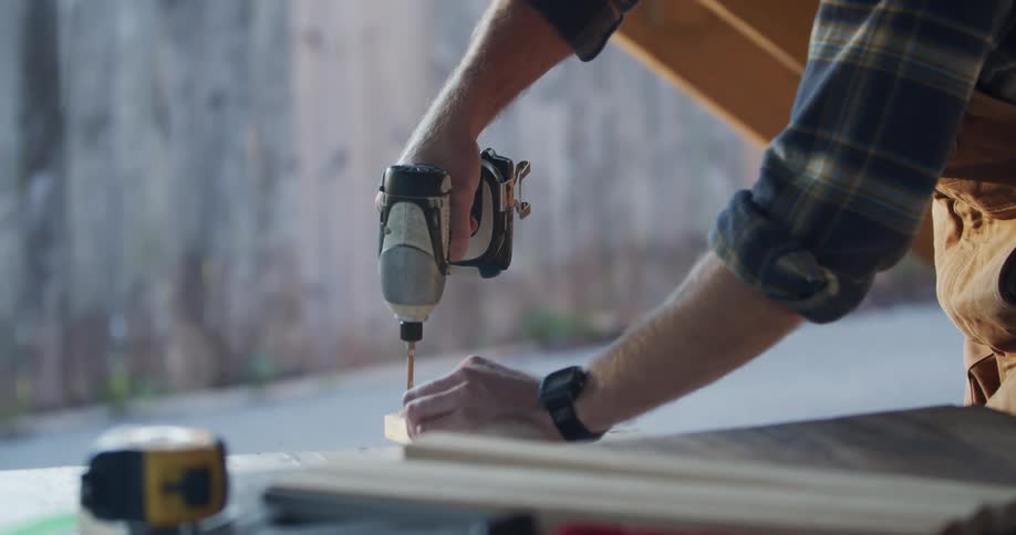 A man in safety goggles uses an electric hand drill to bore a hole into a piece of wood