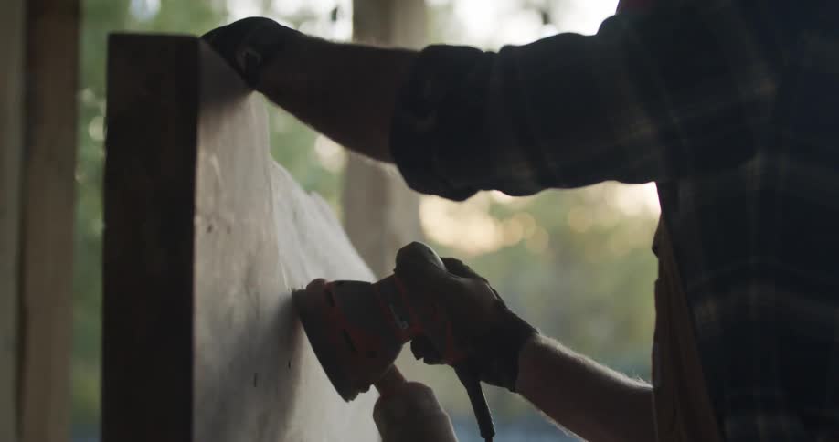 Carpenter uses a power tool to sand a piece of wood outdoors at evening