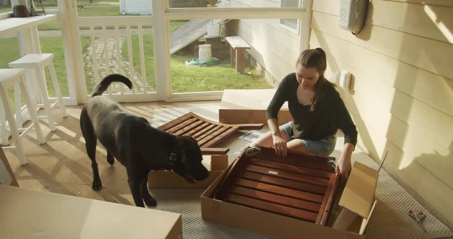A woman in a porch with a dog taking pieces of a furniture out of a box