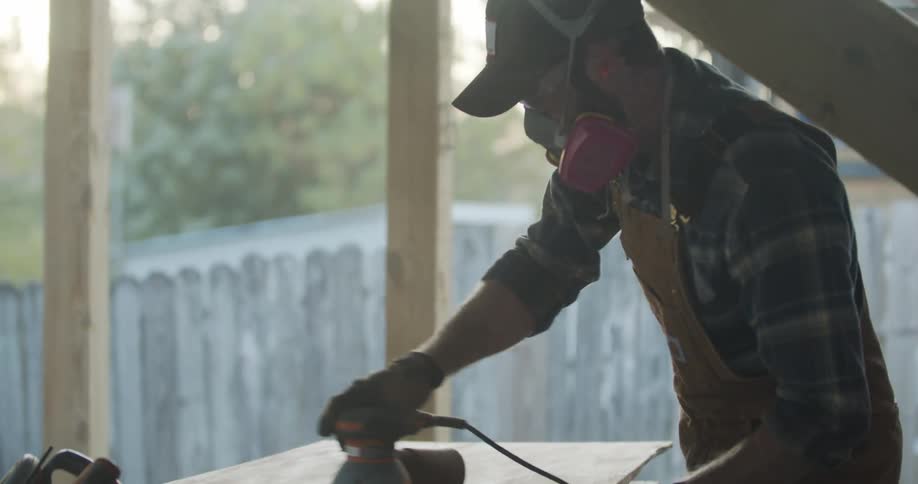 Man sands down wood in to build a structure with power tools on a sunny day with a fence in the background