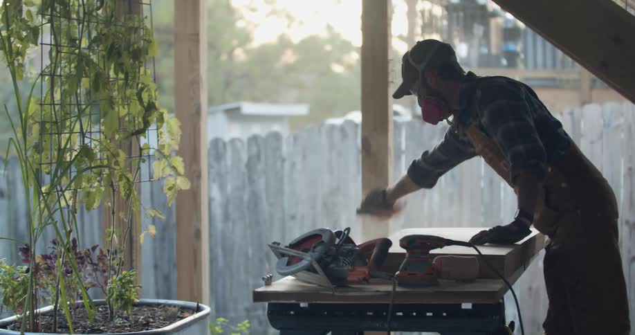 A man wearing a respirator mask brushes saw dust off of a sheet of wood