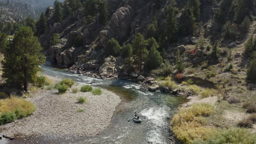 Two people in a raft with paddles going down a river with trees and cliffs in the background