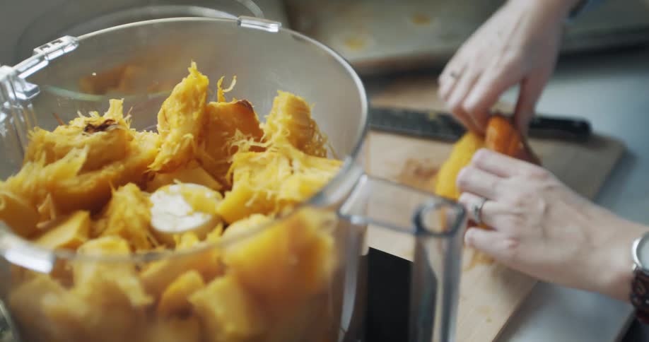 A woman stands at a kitchen counter preparing pumpkin on a cutting board with a food processor sitting to the side