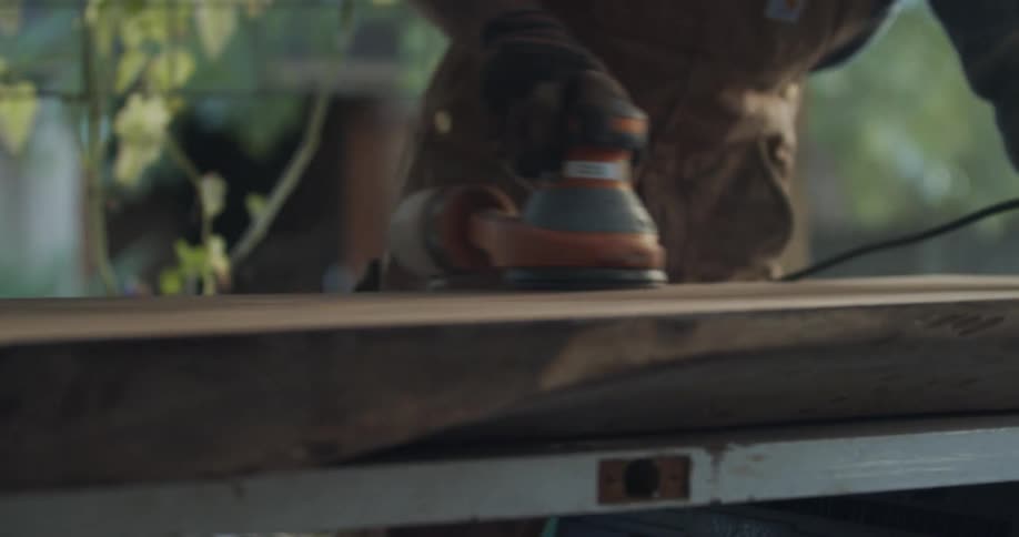 A man wearing brown safety gloves used a power tool on a piece of wood on a table outside