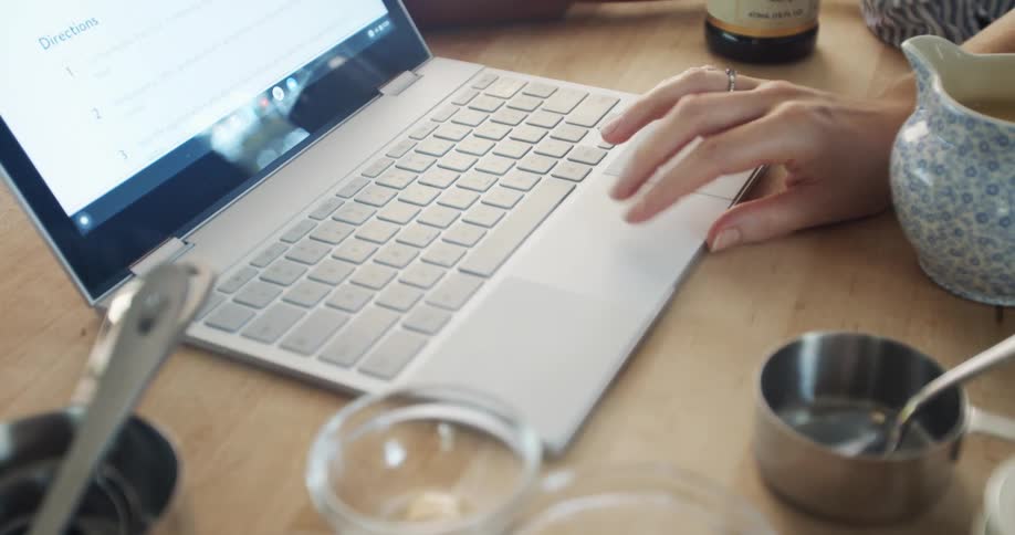 A woman uses her hand to scroll through a document on her laptop while cooking in a kitchen