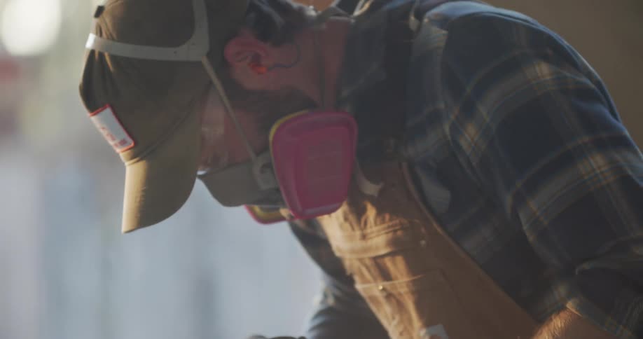 A man uses a power sander on a large piece of wood wearing face protection