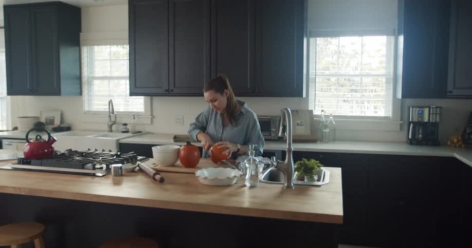 Woman with ponytail and blue shirt cuts pumpkin with knife in the kitchen